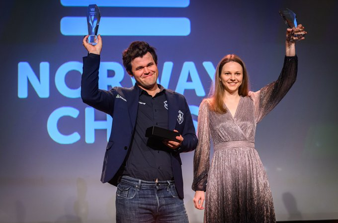 Magnus Carlsen and Anna Muzychuk proudly holding their trophies after winning Norway Chess 2025, standing against a backdrop featuring the tournament logo and Norwegian flags.